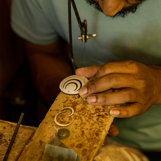 A close-up of a jeweler's hands using a jeweler's saw to cut a small silver ring component on a workbench.