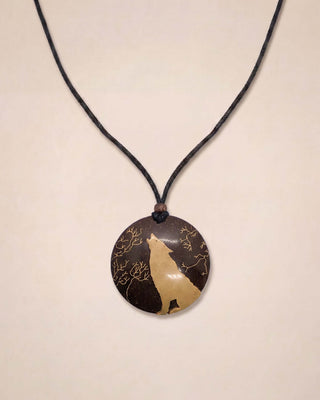 A circular pendant necklace featuring a dark brown and beige wolf silhouette design with gold branch details, suspended on a black cord against a light beige background.
